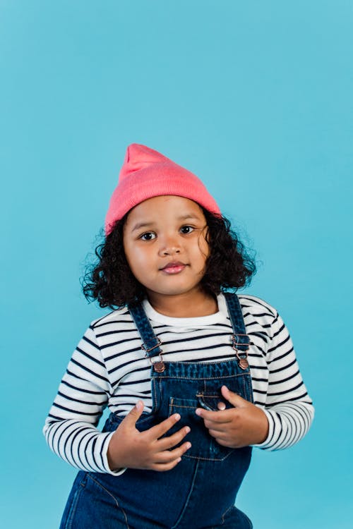 Free Tranquil black little girl with dark curly hair in trendy clothes standing in studio with blue background and looking at camera Stock Photo