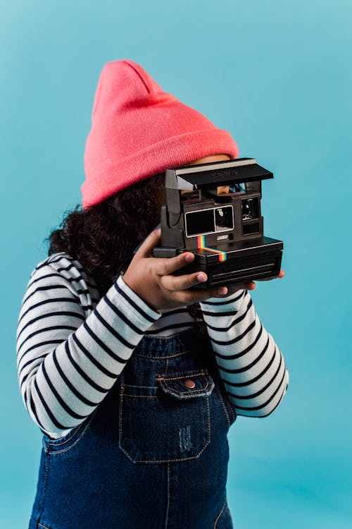 Anonymous ethnic little girl with curly hair in casual clothes taking pictures on instant photo camera against blue background