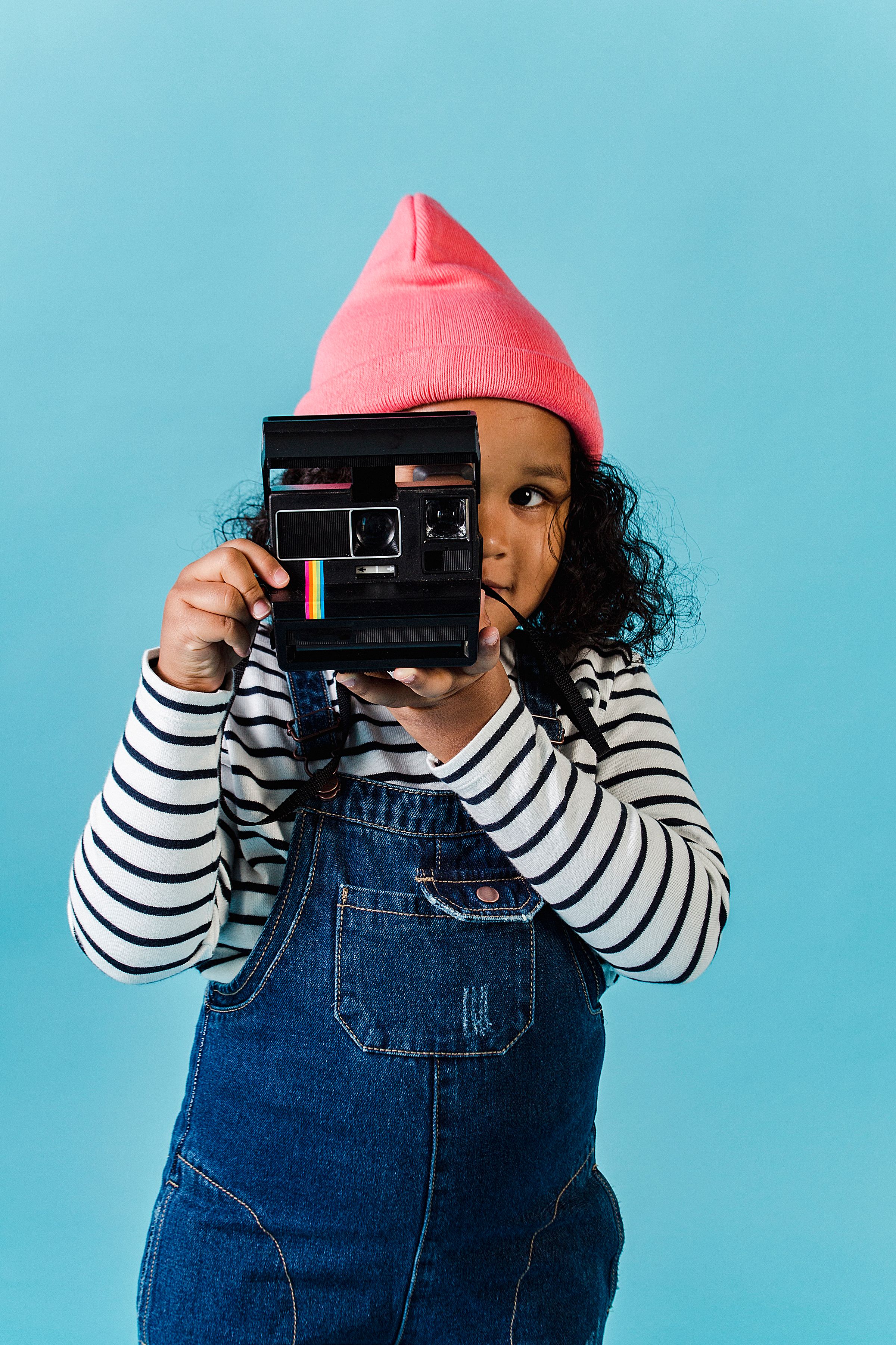 Cheerful child holding a vintage camera wearing a pink hat against a blue background.