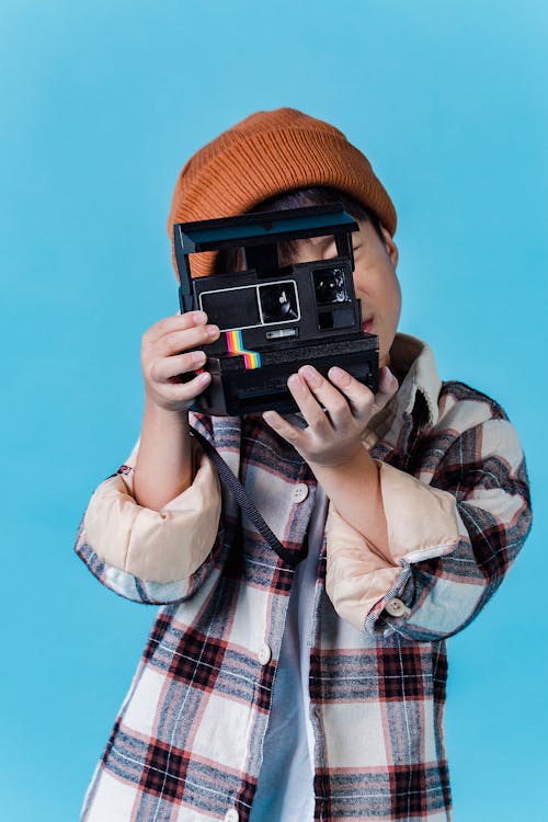 Anonymous Asian boy in stylish clothes standing in studio with blue walls and taking photo on instant photo camera