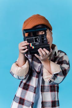 Anonymous Asian boy in stylish clothes standing in studio with blue walls and taking photo on instant photo camera