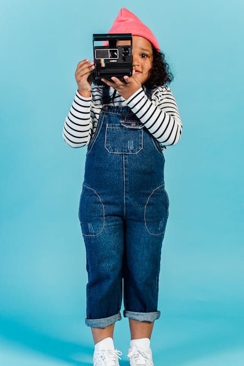 Full body of stylish black girl with curly hair standing in studio with blue wall and taking photo on instant photo camera