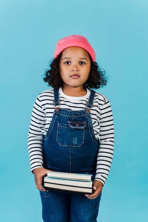 Pensive African American girl with black curly hair in trendy clothes standing with books in hands in studio with blue walls and looking at camera