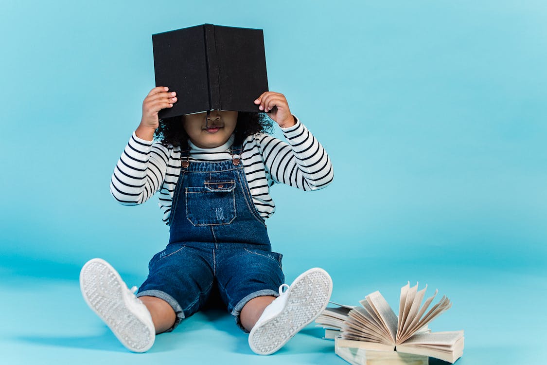 Unrecognizable little African American girl in trendy clothes sitting on floor with textbooks and covering face with book against blue background