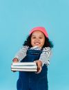 Smiling African American kid wearing hat looking at camera while standing in studio on blue background with books in outstretched arms