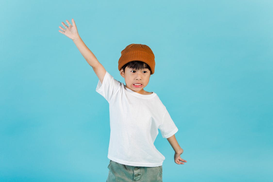 Astonished stylish Asian boy wearing hat and white t shirt looking away while standing with outstretched arms in light studio