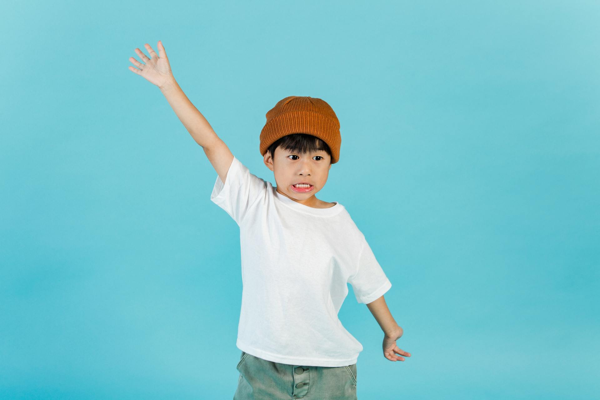 Astonished stylish Asian boy wearing hat and white t shirt looking away while standing with outstretched arms in light studio