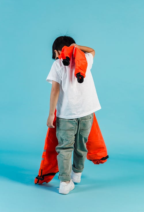 Full body back view of anonymous kid with orange cosmonaut suit standing on blue background in modern studio with hand behind head