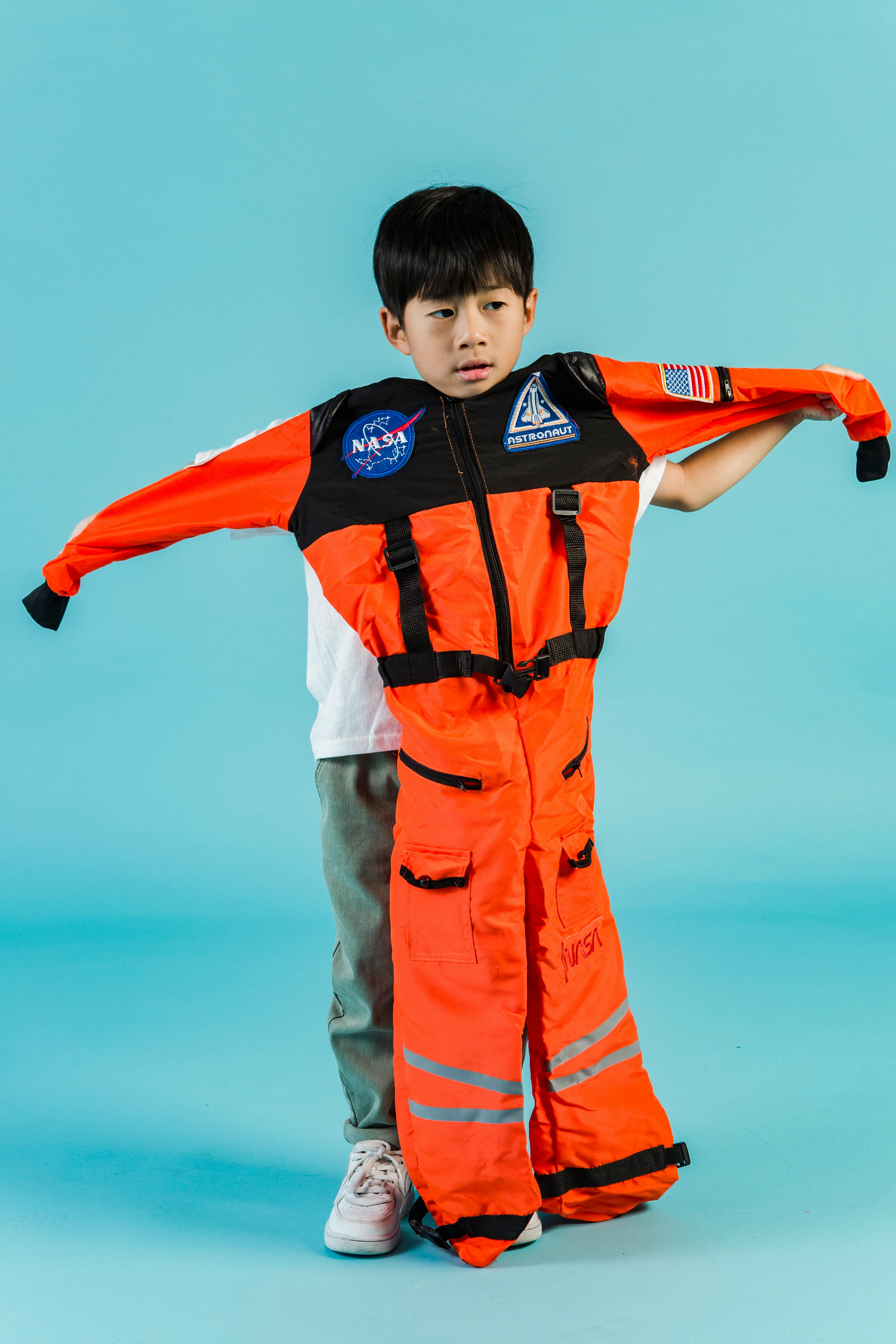 Full body of calm Asian boy with outstretched arms looking away while standing on blue background with orange astronaut suit