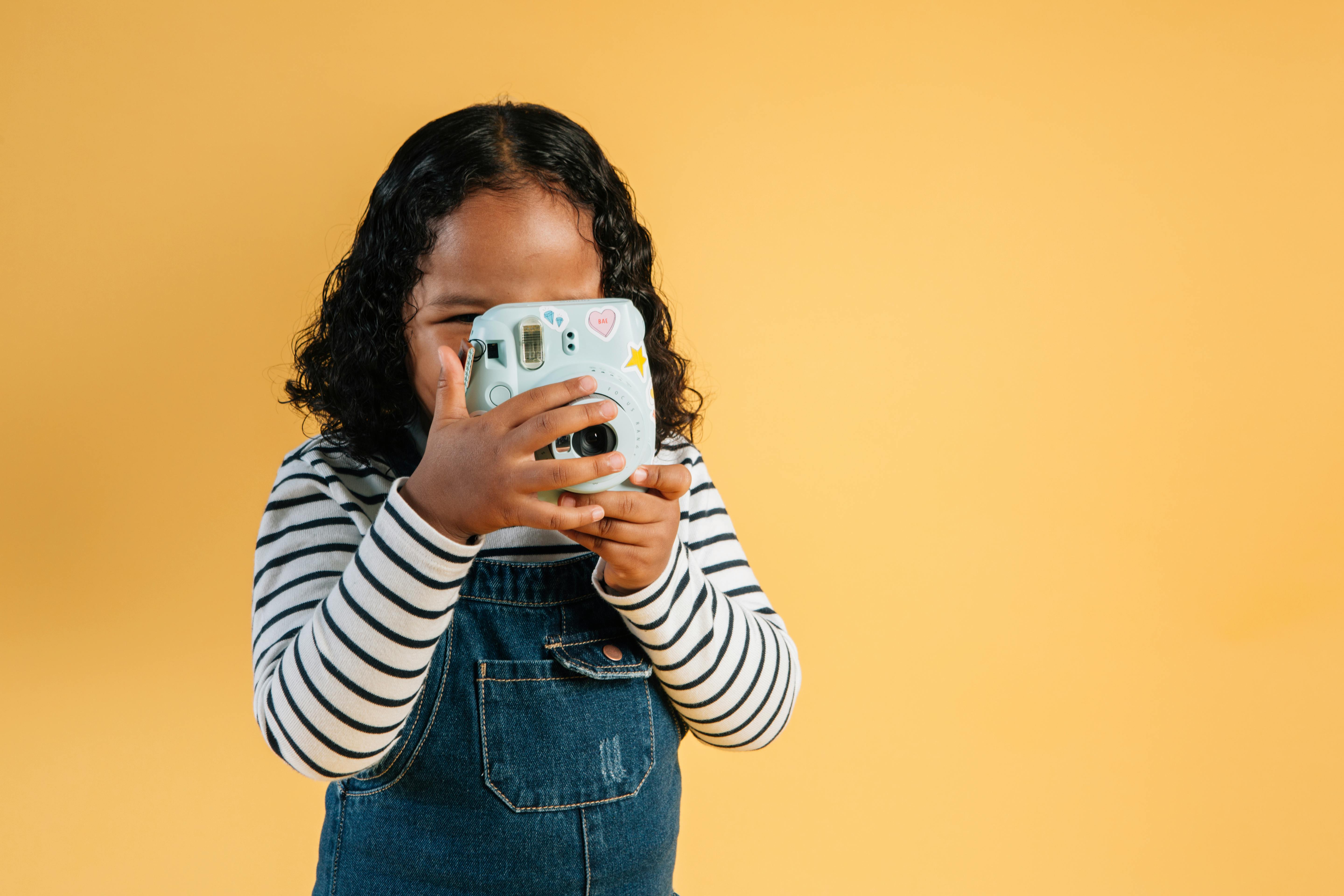 Unrecognizable little African American kid wearing overall standing with instant photo camera in hands on yellow background in modern studio