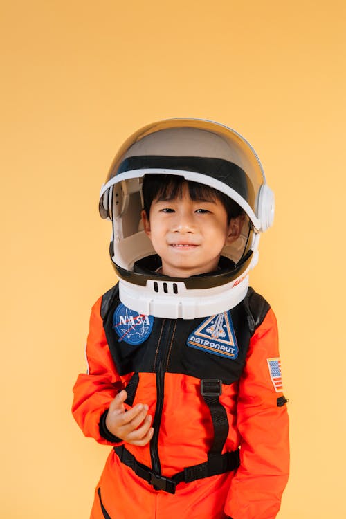 Cheerful Asian kid wearing helmet and orange astronaut suit looking at camera while standing on yellow background in modern studio