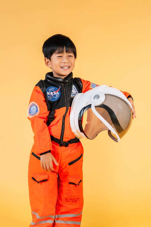 Cheerful Asian kid wearing orange astronaut suit smiling and looking away while standing on yellow background with helmet in hands