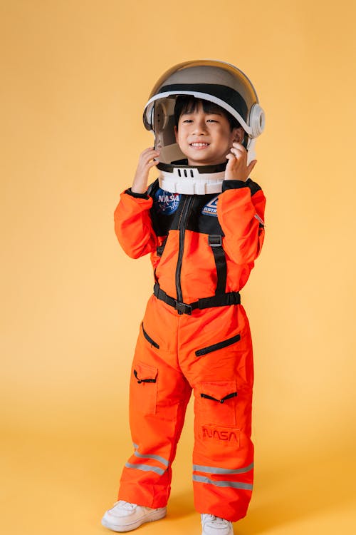 Positive Asian kid wearing orange astronaut costume and helmet smiling and looking away while standing on yellow background in studio