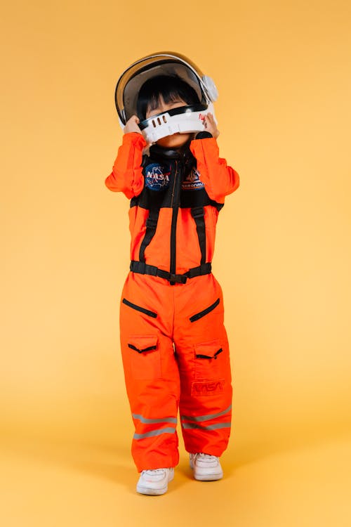 Full body of unrecognizable kid wearing orange cosmonaut suit taking off helmet while standing on yellow background in modern studio