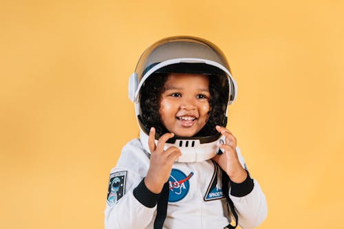 Delighted little African American girl in white space suit touching helmet while looking away on orange background
