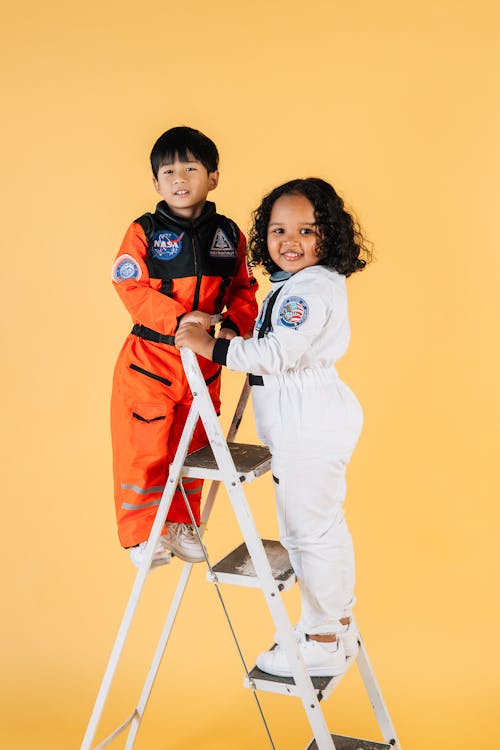 Smiling multiracial children in astronaut costumes standing on ladder and looking away against yellow background in studio