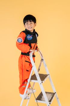 一个快乐的孩子穿着橙色太空服，在工作室的黄色背景前爬上梯子