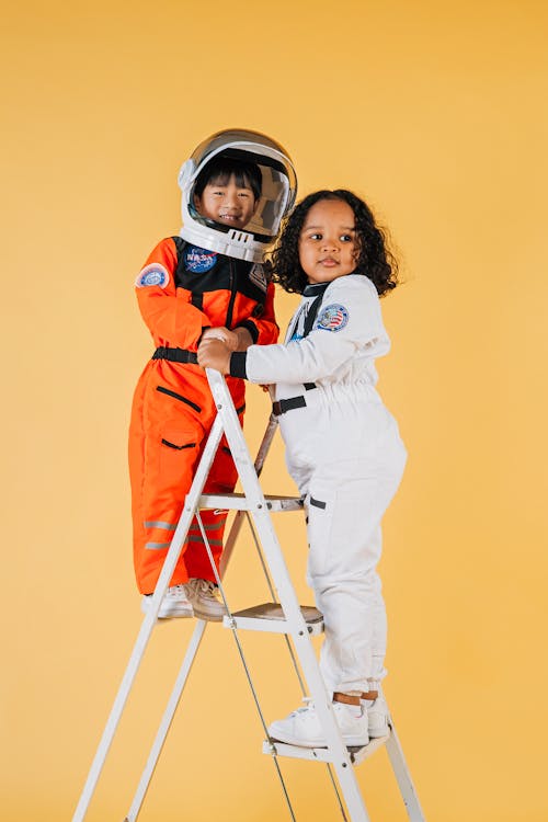 Full length of positive multiethnic children in colorful astronaut costumes standing on top of ladder against yellow background in studio