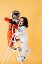 Cheerful multiracial children in astronaut costumes standing on ladder in studio against yellow background and looking away