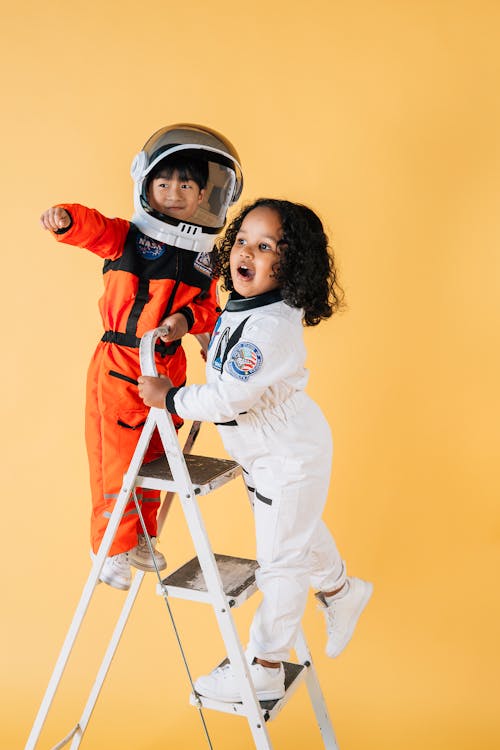 Free Cheerful multiracial children in astronaut costumes standing on ladder in studio against yellow background and looking away Stock Photo
