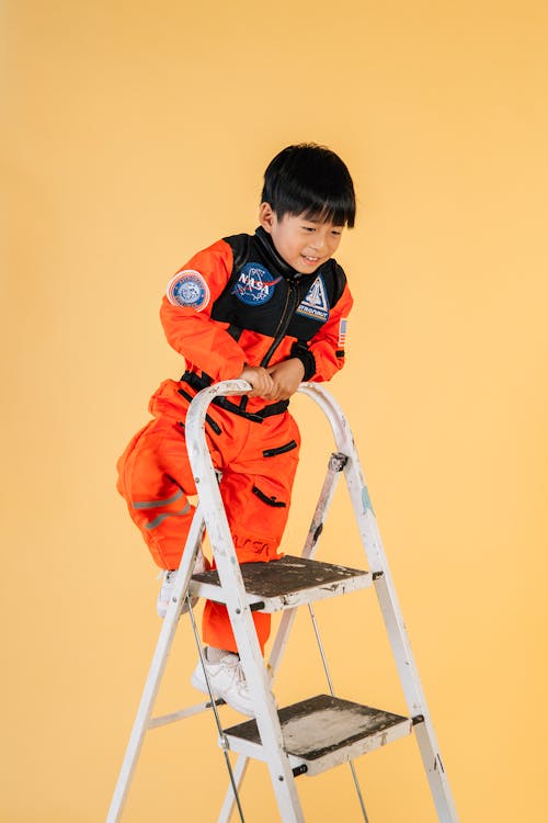 Playful Asian boy in orange astronaut costume climbing up ladder in studio against yellow background