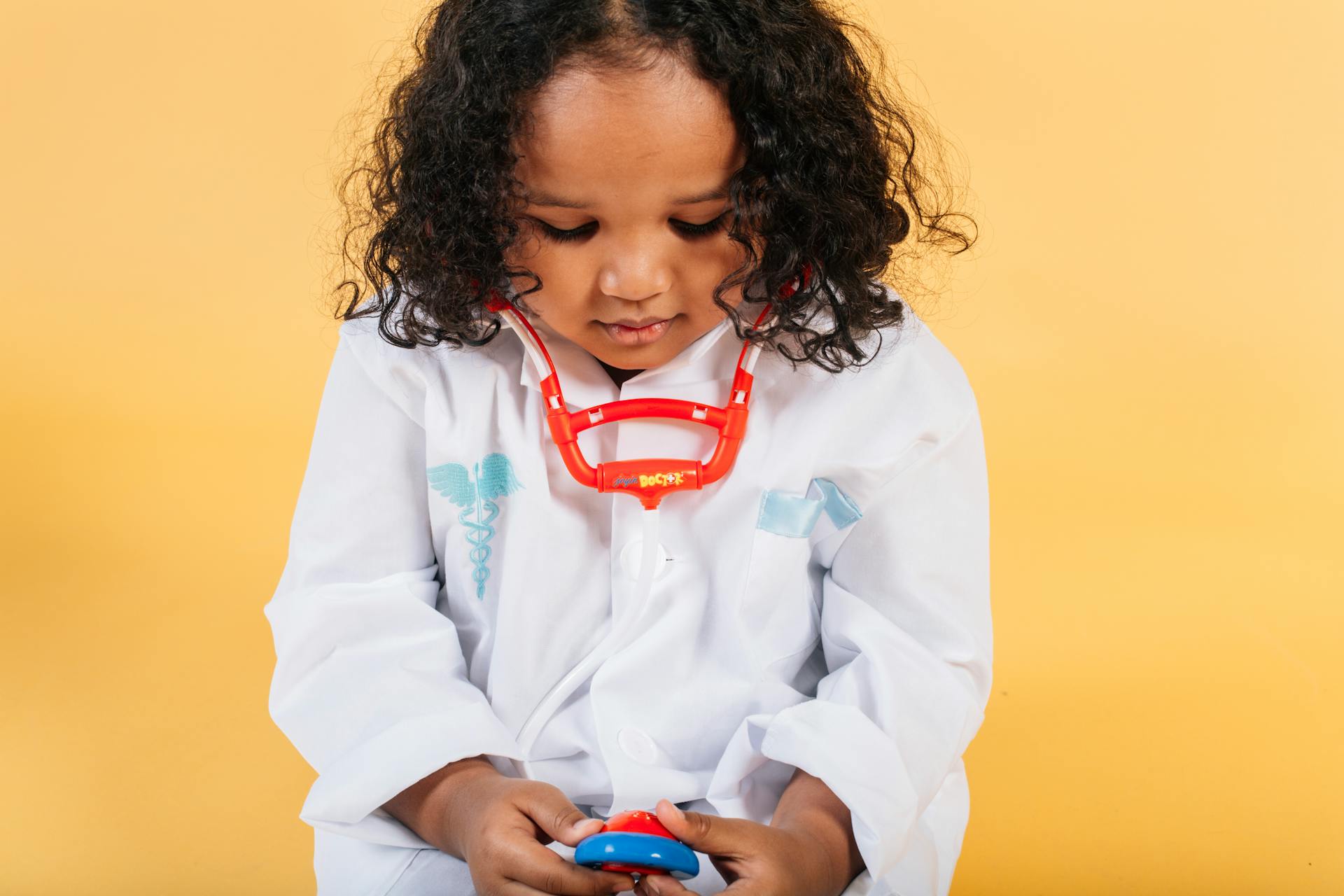 Tranquil little African American girl with curly hair in medical costume and stethoscope looking down against yellow background in studio