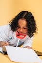 Focused curly African American girl in doctor costume with toy stethoscope taking notes in medical card while lying on floor in studio on yellow background