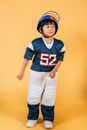 Full body focused ethnic kid in American football player uniform with number and helmet standing in studio on yellow background and looking away