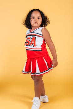 Serious curly black girl in cheerleader costume and sneakers standing in confident pose with round chest on yellow background in studio