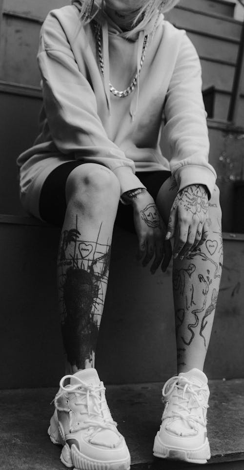 Free Grayscale Photo of a Person in Sweatshirt with Tattoos on Hands and Legs Stock Photo