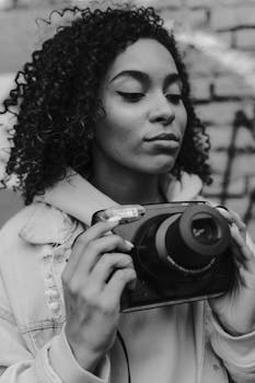 Black and white portrait of a young woman with curly hair holding a vintage camera against a graffiti wall.