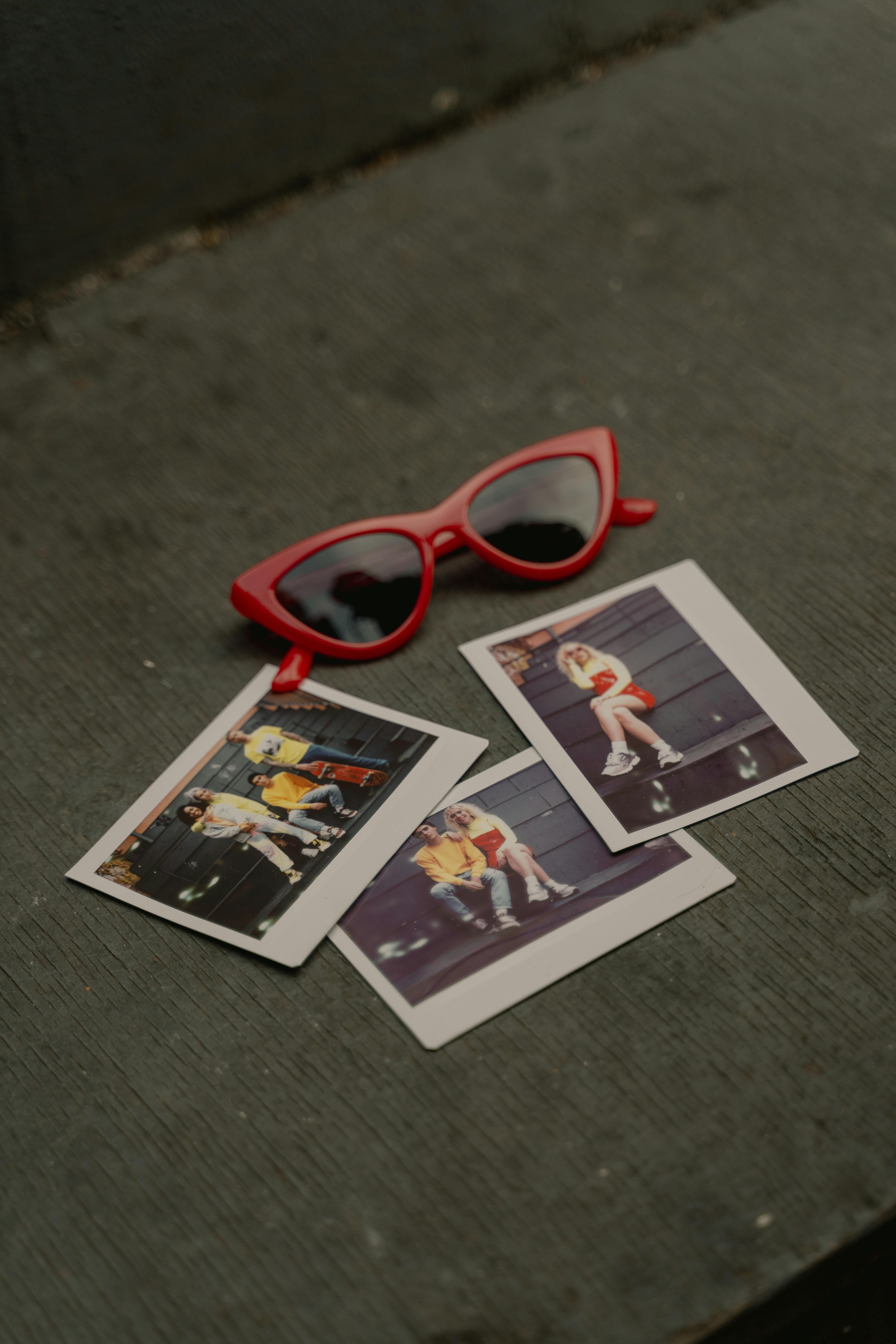 Close-up of stylish red sunglasses next to nostalgic polaroid photos on a wooden surface.
