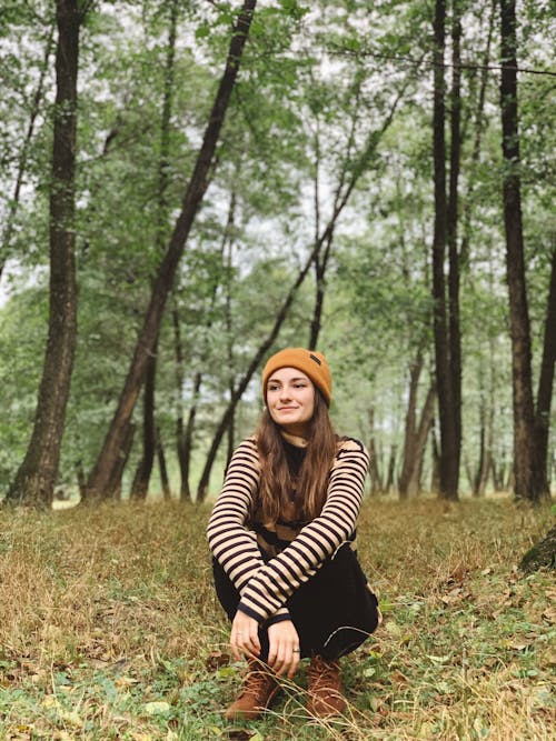 Full body of optimistic young female in casual clothes and hat resting on dry grassy ground in forest on autumn day