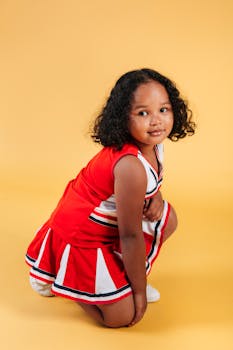A young girl wearing a vibrant cheerleader outfit poses confidently on a yellow backdrop.