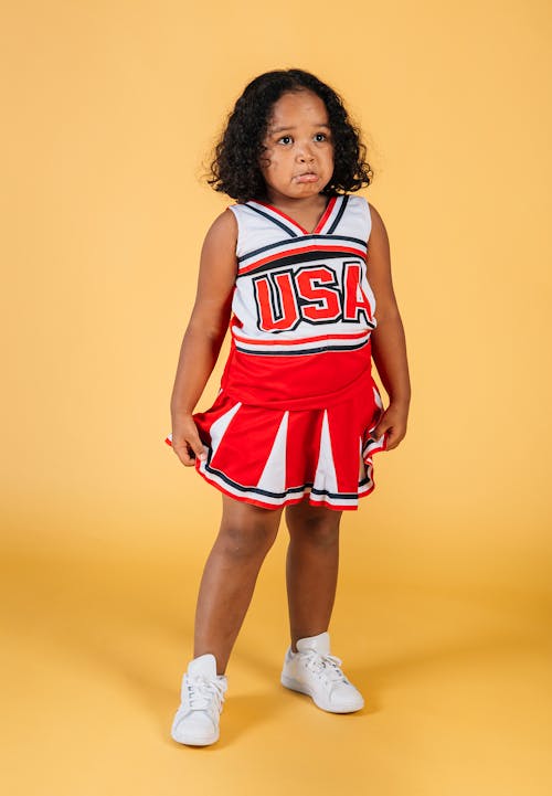 Full body African American curly female kid in cheerleader costume standing on yellow background with offended face in studio and looking away