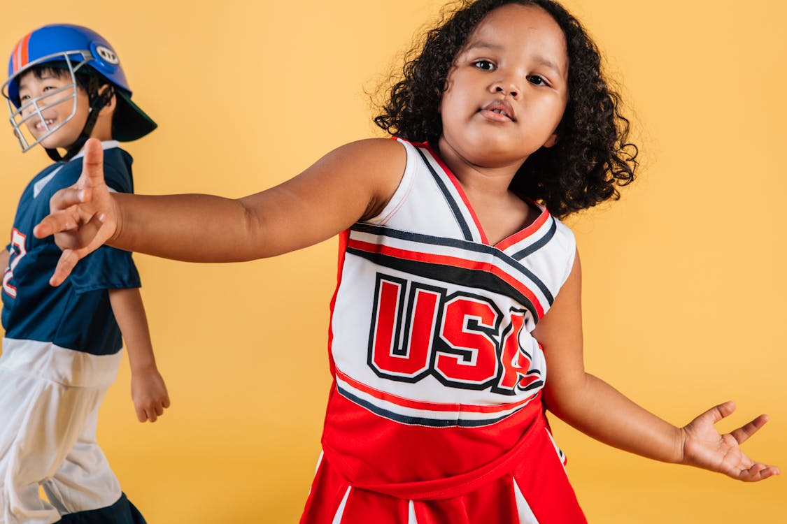 Cute African American girl wearing cheerleader uniform dancing on yellow background near cheerful Asian boy in helmet and football player costume