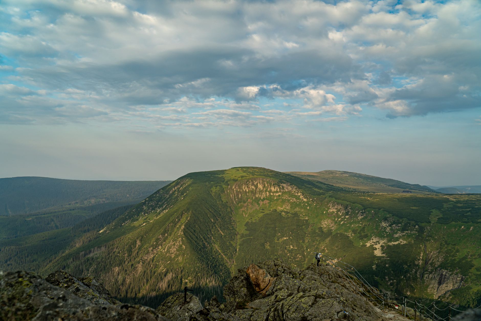 A hiker standing on a rocky peak with lush green mountains under a cloudy sky. — free online jigsaw puzzle on JigsawPuzzlesFreeOnline.org