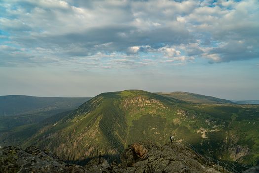 A hiker standing on a rocky peak with lush green mountains under a cloudy sky., free online jigsaw puzzle on JigsawPuzzlesFreeOnline.org