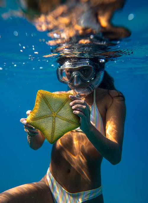 Young female diver in mask and swimsuit holding yellow starfish while looking at camera