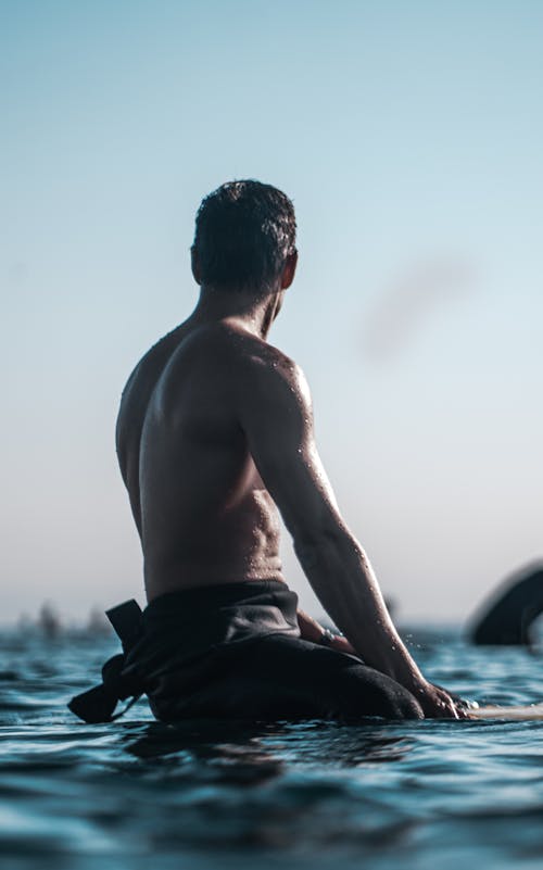 Back view of shirtless male with muscular torso sitting on surfboard and looking at sky