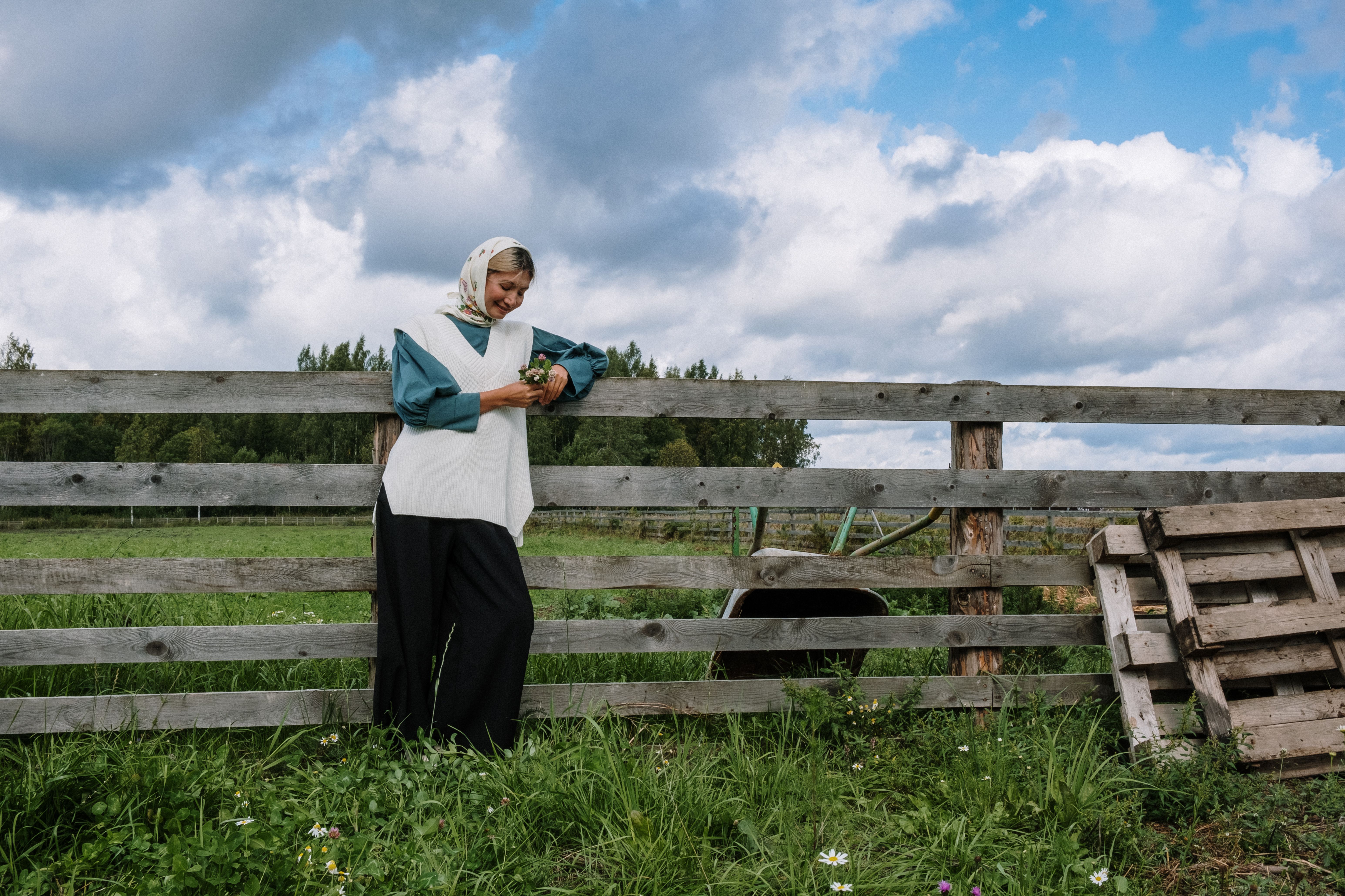 A woman in a headscarf stands by a wooden fence in a rural setting.