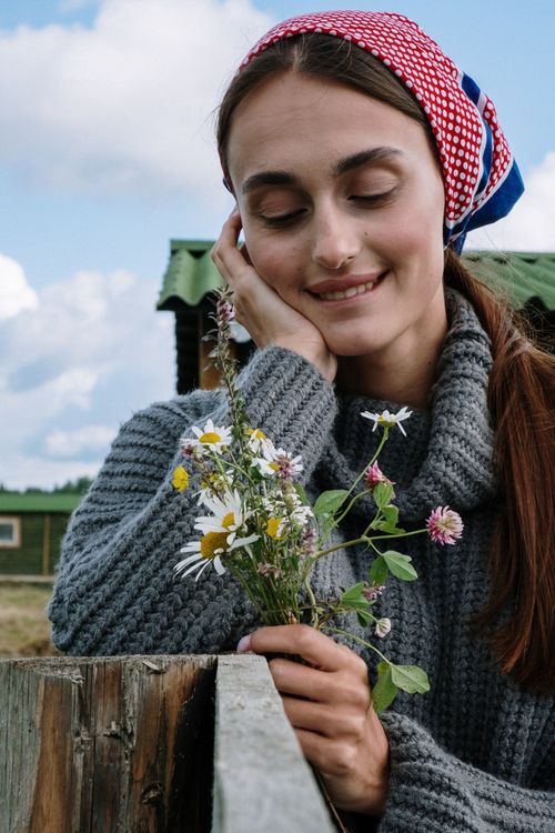 Free Smiling woman in a sweater and headscarf holds wildflowers outdoors in a rural setting. Stock Photo