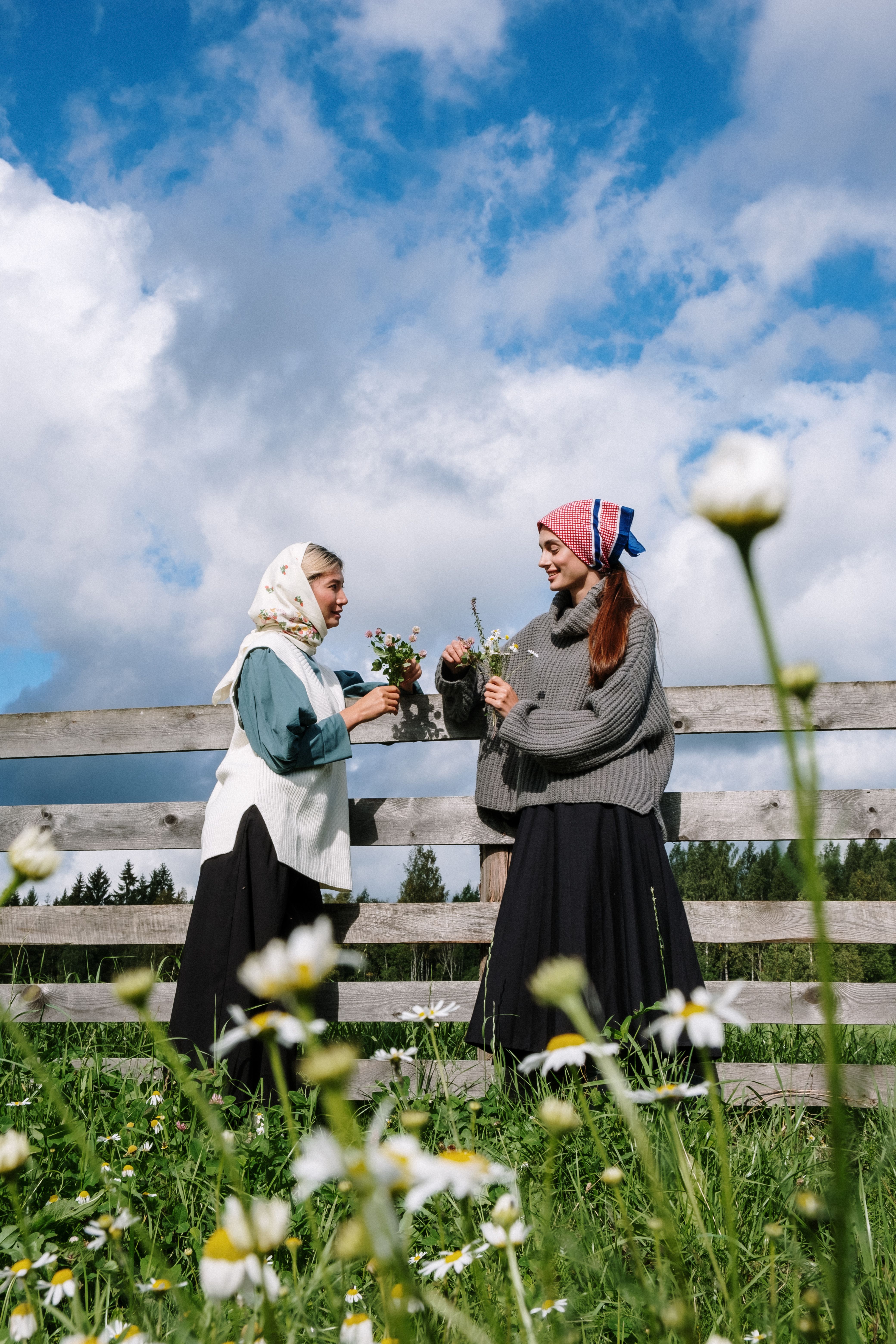 Two women in headscarves gather flowers by a rustic wooden fence on a sunny day.