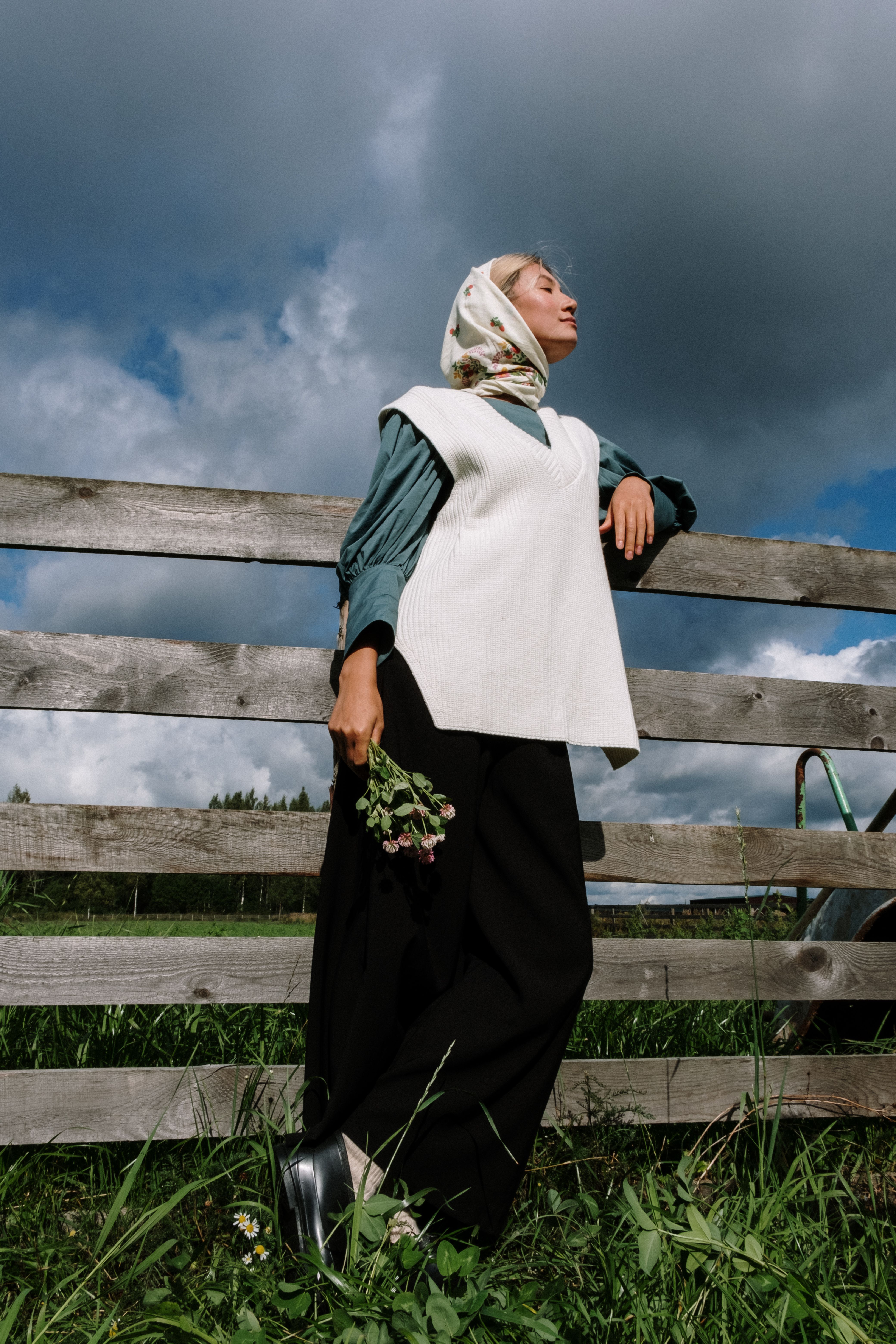 A young woman in a headscarf stands by a wooden fence under a dramatic sky in the countryside.