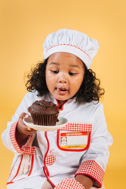 Excited African American girl wearing chef uniform and hat sitting on yellow background and licking lips while looking at tasty chocolate muffin