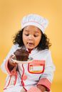 Excited African American girl wearing chef uniform and hat sitting on yellow background and licking lips while looking at tasty chocolate muffin