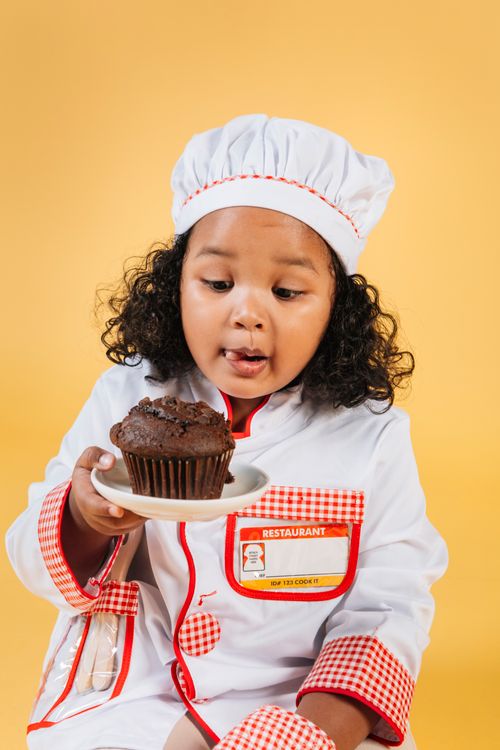 Free Excited African American girl wearing chef uniform and hat sitting on yellow background and licking lips while looking at tasty chocolate muffin Stock Photo