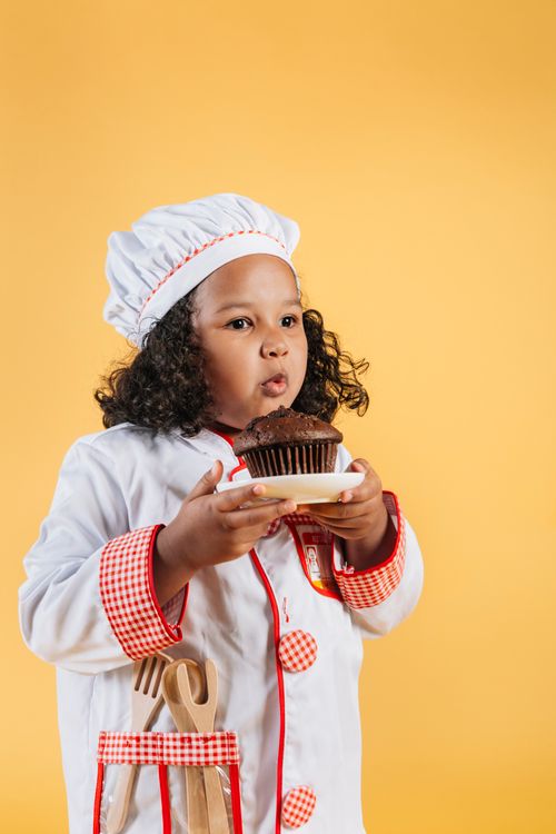 Free Cute little African American girl wearing chef uniform blowing on delicious chocolate cake in hands against brown background Stock Photo