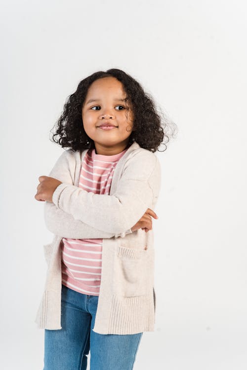 Cheerful little African American girl in jeans and knitted cardigan standing with arms crossed and looking away dreamy against white background