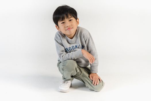 Content little Asian boy in stylish outfit sitting on haunches and looking at camera confidently during photo shoot in light studio